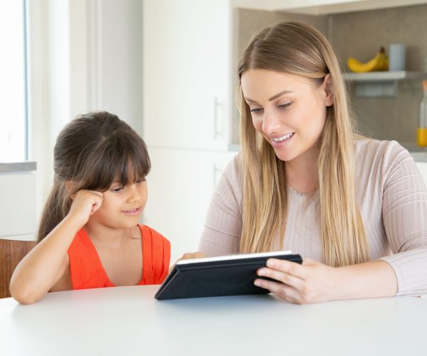 Blonde mom holding tablet and showing it to daughter. Cute girl sitting at kitchen table with young beautiful mother and smiling. Family time, digital technology, childhood and weekend concept